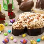 Itens de Páscoa sobre uma mesa clara: há um bolo com cobertura bege, cercado por pequenos ovos de páscoa embalados e coelhos no formato de chocolate