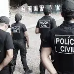 Integrantes da Polícia Civil, vestidos de roupa preta, aparecem de costas para a câmera, enquanto olham para um muro