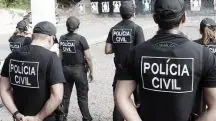 Integrantes da Polícia Civil, vestidos de roupa preta, aparecem de costas para a câmera, enquanto olham para um muro