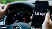 Imagem de um motorista, dentro de um carro, com a mão esquerda no volante e a direita segundo um celular, que mostra o aplicativo da Uber ligado