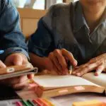 Imagem mostra duas pessoas à mesa, mas sem exibir os rostos, à mesa em momento de leitura