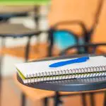 Caderno escolar em cima de uma mesa de estudo preta. As folhas são brancas, e há uma caneta de cor azul em cima do caderno