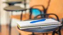 Caderno escolar em cima de uma mesa de estudo preta. As folhas são brancas, e há uma caneta de cor azul em cima do caderno