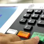 Urna eletrônica brasileira com tela azul exibindo a palavra "FIM" e teclado numérico, o dedo de um eleitor está prestes a pressionar o botão 'CONFIRMA', verde, para finalizar o voto nas eleições municipais
