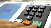 Urna eletrônica brasileira com tela azul exibindo a palavra "FIM" e teclado numérico, o dedo de um eleitor está prestes a pressionar o botão 'CONFIRMA', verde, para finalizar o voto nas eleições municipais