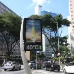 Um termômetro gigante fincado no chão do centro de São Paulo, com prédios dos lados e o céu aberto, com algumas nuvens