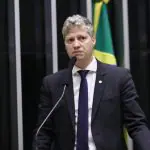 deputado Marcel van Hattem (Novo) usa terno, camisa e grava em palanque. Atrás, há uma bandeira do Brasil