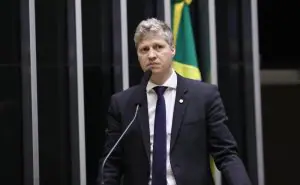 deputado Marcel van Hattem (Novo) usa terno, camisa e grava em palanque. Atrás, há uma bandeira do Brasil