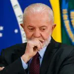 Lula, com a mão direita no rosto; ele está no Palácio do Planalto, em Brasília, usando paletó preto, gravata vinho e camisa azul claro; ele está com semblante de alguém sério, pensativo; atrás dele, há a bandeira do Brasil