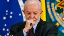 Lula, com a mão direita no rosto; ele está no Palácio do Planalto, em Brasília, usando paletó preto, gravata vinho e camisa azul claro; ele está com semblante de alguém sério, pensativo; atrás dele, há a bandeira do Brasil