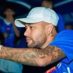 Neymar usa boné branco, camisa azul e segura celular com a mão esquerda