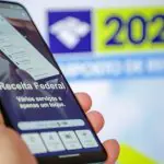 Imagem mostra uma mão segurando um aparelho de telefone celular, que está com o aplicativo da Receita Federal aberto