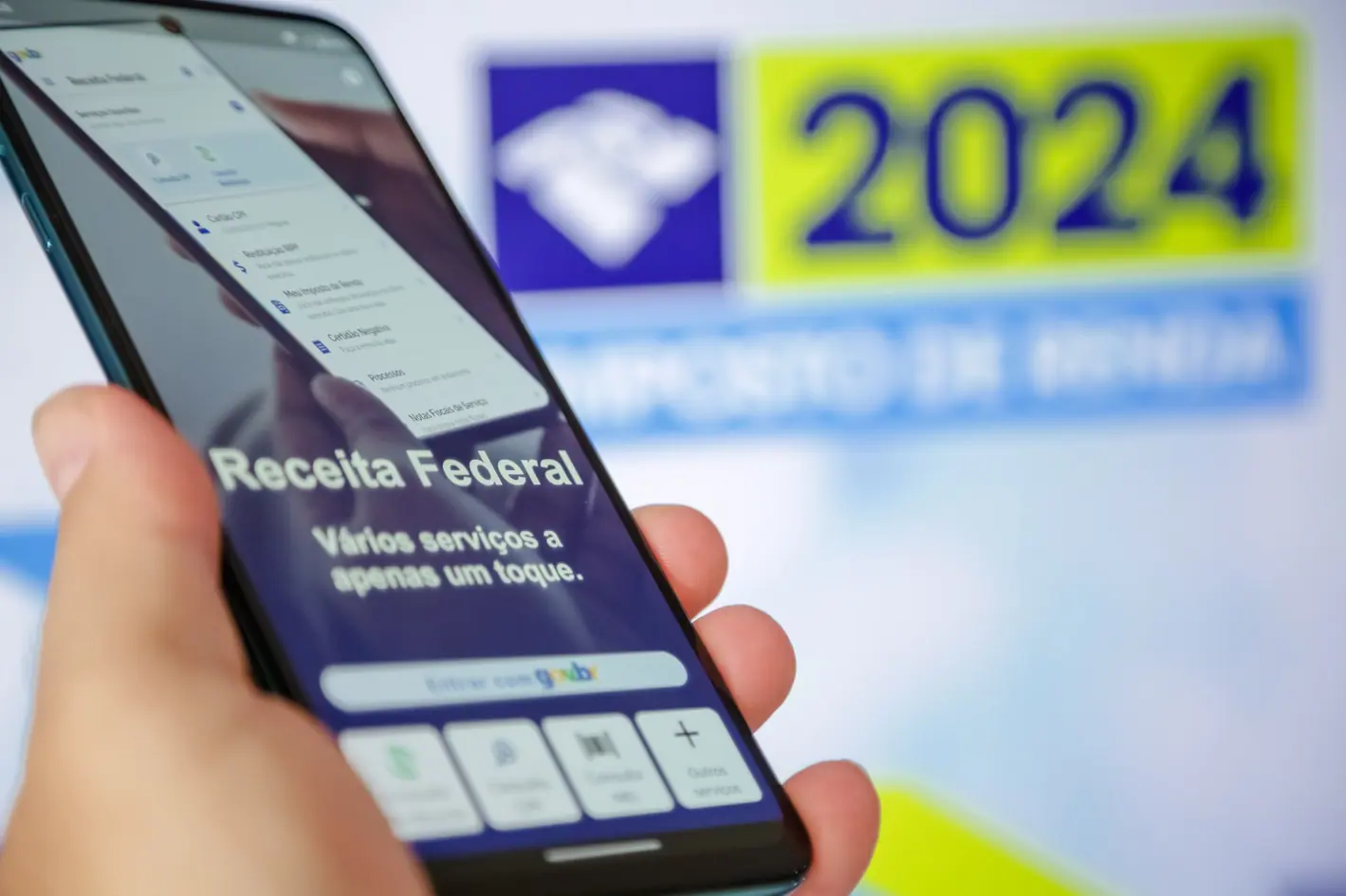 Imagem mostra uma mão segurando um aparelho de telefone celular, que está com o aplicativo da Receita Federal aberto