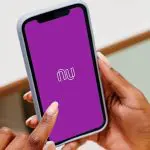 Foto mostra mãos femininas segurando celular. Na tela, aparece o logo do Nubank