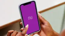 Foto mostra mãos femininas segurando celular. Na tela, aparece o logo do Nubank