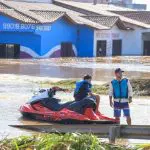 Imagem de bairro alagado no município de Eldorado do Sul (RS); homem em pé aparece de colete salva-vida, há um garoto sentado sobre um jet ski em rua alagada