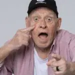 Foto mostra o narrador Silvio Luiz. Ele usa uma camisa rosa e um boné preto
