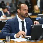 Eduardo Bolsonaro, deputado federal pelo PL, está sentado, com um computador em sua frente. Ele fala ao microfone do Legislativo e usa terno, camisa e gravata