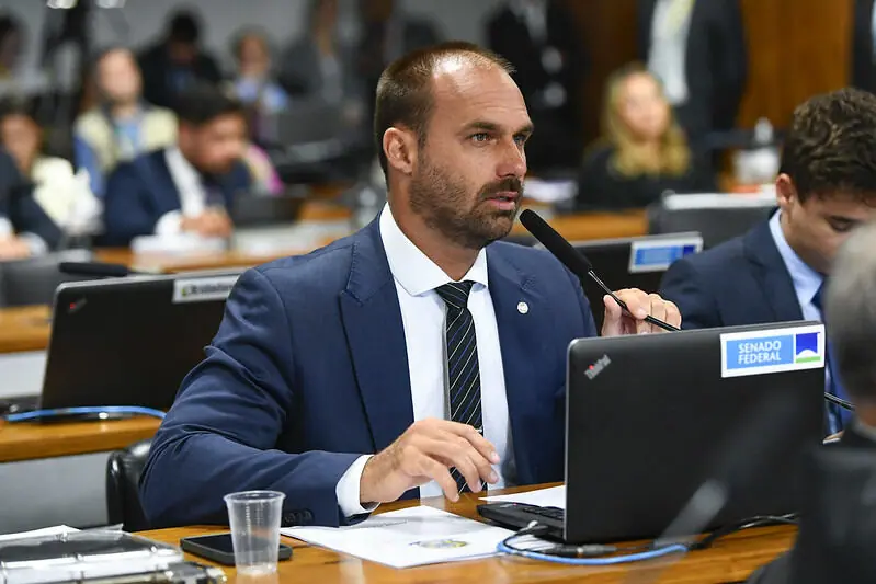 Eduardo Bolsonaro, deputado federal pelo PL, está sentado, com um computador em sua frente. Ele fala ao microfone do Legislativo e usa terno, camisa e gravata