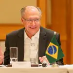 Imagem do vice-presidente Geraldo Alckmin, vestindo blazer preto e camisa social branca, sentado em frente a bandeira do Brasil pequena