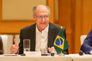 Imagem do vice-presidente Geraldo Alckmin, vestindo blazer preto e camisa social branca, sentado em frente a bandeira do Brasil pequena