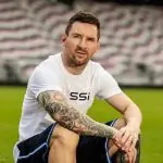 Lionel Messi veste camiseta branca e shorts preto, sentado no meio do campo de futebol