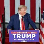 Donald Trump, candidato às eleições dos EUA, Estados Unidos, aparece em comício, em frente a púlpito e microfone. Ao fundo, há bandeiras dos Estados Unidos, e ele usa terno, camisa e gravata