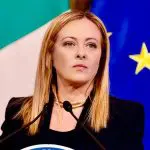 Giorgia Meloni critica acordo para cargos da União Europeia. Ela aparece em frente a púlpito, com bandeiras da Itália e da União Europeia ao fundo