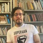 Jean Wyllys aparece com camisa branca e rosa, onde está escrito Super Man. Ao fundo, há uma estante cheia de livros