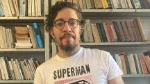 Jean Wyllys aparece com camisa branca e rosa, onde está escrito Super Man. Ao fundo, há uma estante cheia de livros