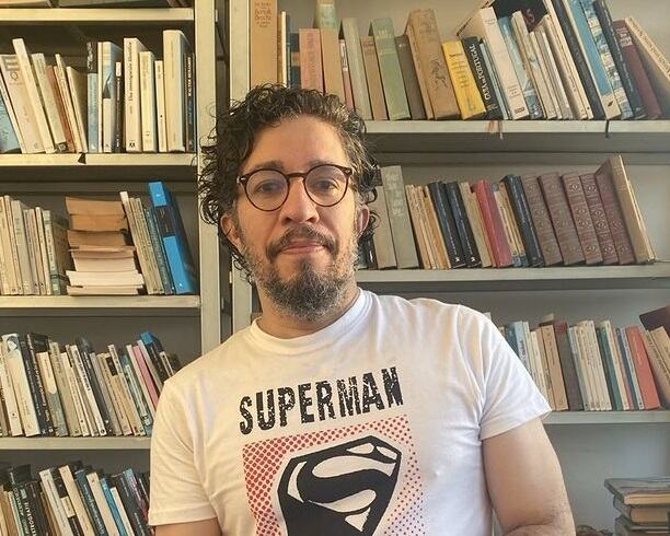 Jean Wyllys aparece com camisa branca e rosa, onde está escrito Super Man. Ao fundo, há uma estante cheia de livros