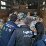 Três agentes do Ministério Público do RS estão de costas para a foto, com colete azul, fiscalizando um caminhão com doações às vítimas das enchentes, supostamente desviadas
