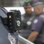 Imagem mostra policial militar usando câmera e sua farda. Ao fundo, aparecem outros funcionários da Polícia Militar