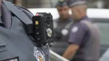 Imagem mostra policial militar usando câmera e sua farda. Ao fundo, aparecem outros funcionários da Polícia Militar