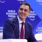 Primeiro-ministro da Espanha, Pedro Sánchez, vestindo terno azul, camisa social branca e gravata vermelha. Ele está em frente a um púlpito e a um microfone. Ao fundo, há um grande painel azul
