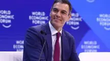 Primeiro-ministro da Espanha, Pedro Sánchez, vestindo terno azul, camisa social branca e gravata vermelha. Ele está em frente a um púlpito e a um microfone. Ao fundo, há um grande painel azul