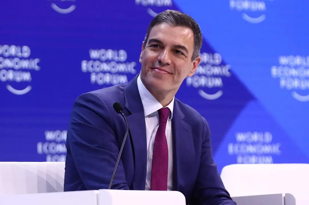 Primeiro-ministro da Espanha, Pedro Sánchez, vestindo terno azul, camisa social branca e gravata vermelha. Ele está em frente a um púlpito e a um microfone. Ao fundo, há um grande painel azul