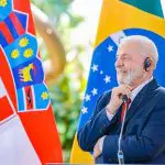 Presidente Lula, de terno, em frente à um púlpito, sorri e olha para o lado. Atrás dele, há bandeiras do Brasil e da Croácia