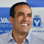 Bruno Reis aparece sorrindo. Ele está de camisa azul claro. Atrás, banner com logo da Prefeitura de Salvador