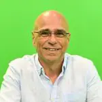 Farid Madi em imagem de divulgação; ele está sorrindo, com visual careca e sem bigode, de óculos e camisa social claro; ao fundo, um painel verde claro