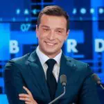 O político francês Jordan Bardella aparece sorridente, com braços cruzados na altura do tórax; ele está de paletó e gravata de cor azul e camisa social branca