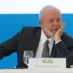 O presidente Luiz Inácio Lula da Silva, em um evento; ele está com feição de desolado, com a mão esquerda em seu rosto e a mão direita sobre a mesa