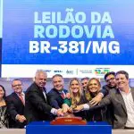 Imagem com cartaz azul com nome da BR-381. Homens de terno e mulheres juntos seguram o martelo em leilão
