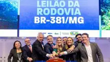 Imagem com cartaz azul com nome da BR-381. Homens de terno e mulheres juntos seguram o martelo em leilão