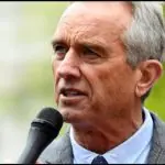 Imagem de Kennedy Jr. com fundo desfocado falando ao microfone