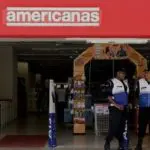 Em frente à fachada das Lojas Americanas, dois seguranças vestindo coletes azul e branco