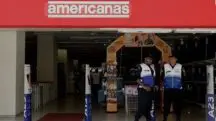 Em frente à fachada das Lojas Americanas, dois seguranças vestindo coletes azul e branco