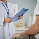 Médica com um paciente em cima da maca; ele está sentado e com as mãos sobre as pernas | Foto: Reprodução/Freepik