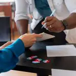 Imagem de homem com caneta assinando cédula e entregando a outro homem de camisa azul. Ao lado bandeira dos EUA.