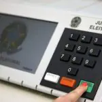 Modelo de urna eletrônica usada no sistema eleitoral brasileiro. Na imagem, mostra-se um dedo indicador direito, possivelmente de uma mulher, pois está com esmalte vermelho na unha, em que simula-se o momento final da votação: teclando o botão verde, de 'confirma'